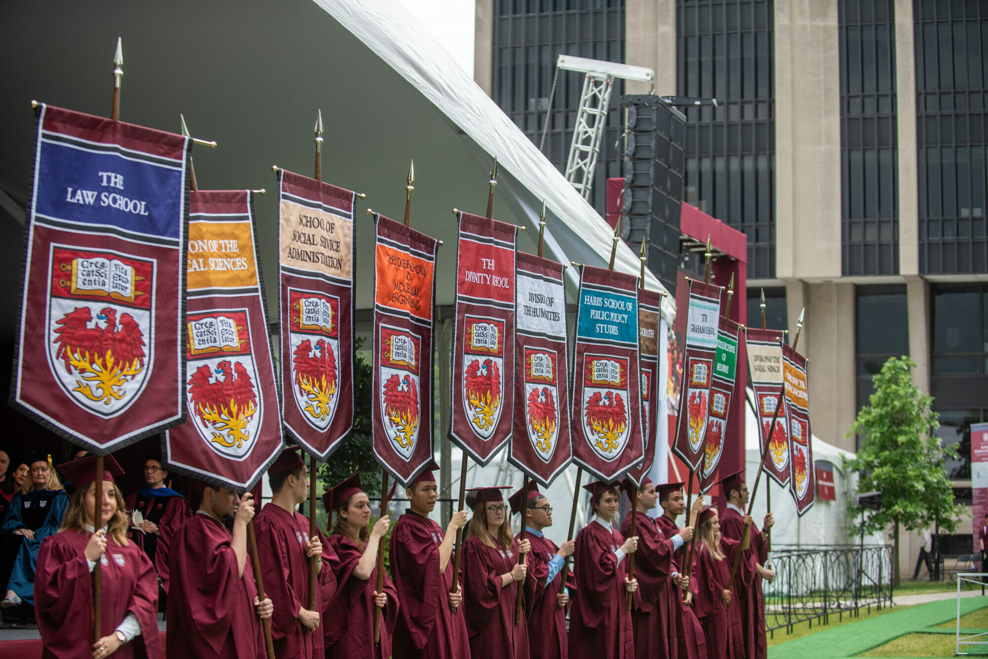Students in graduation robes hold banners representing each graduate school
